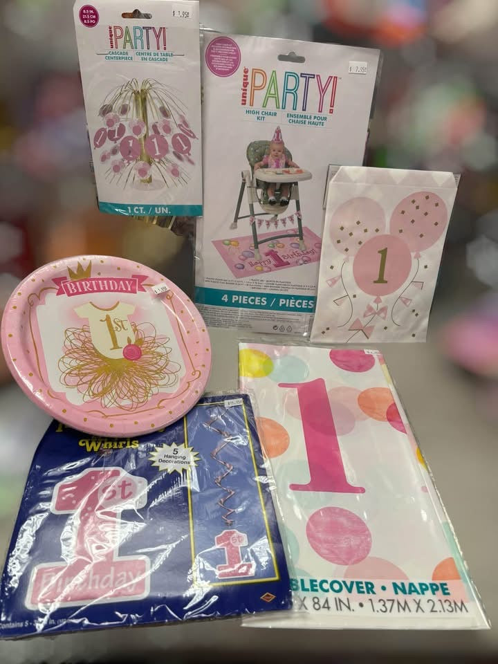 A 1st birthday party decoration kit containing a pink and gold '1st Birthday' plate, a party hat napkin, a balloon print, a high chair mat, and a blue napkin cover, all in retail packaging.