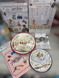A Harry Potter themed party decoration kit displayed with its packaging and contents, including printed plates, napkins, and small accessories on a blurred gray surface.