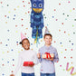 Two children with party hats standing next to a PJ Masks balloon against a polka dot background.