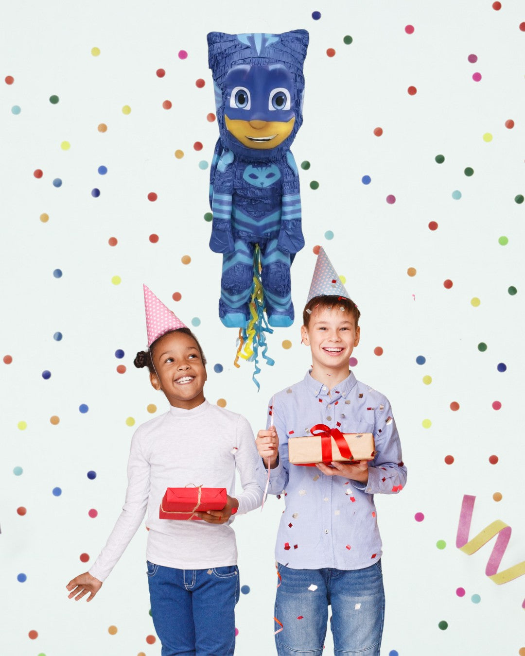 Two children with party hats standing next to a PJ Masks balloon against a polka dot background.